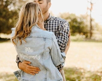 May include: A couple embracing outdoors. The woman is wearing a light blue denim jacket with the text "Mrs. Montgomery" embroidered on the back. The man is wearing a plaid shirt.