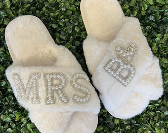 May include: A pair of white fluffy slippers with pearl embellishments. One slipper says "MRS" and the other says "BRIDE".