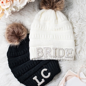 May include: Two knit beanies with faux fur pom poms. One beanie is white with the word "BRIDE" in rhinestones. The other beanie is black with the initials "LC" in rhinestones.