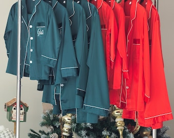 May include: A set of six satin pajamas in green and red with white piping. The pajamas have a button-up front and a collar. The green pajamas have the name "Gia" embroidered on the front. The pajamas are hanging on a rack in front of a Christmas tree.