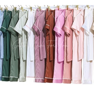 May include: A display of 16 different colored satin pajamas with white piping. The pajamas have short sleeves and a collared top. The pants are long and have a straight leg.