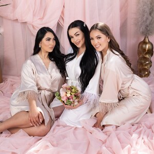 May include: Three women wearing satin robes with feather trim. The robes are in shades of white and blush pink. The women are smiling and looking at the camera. One woman is holding a bouquet of flowers.