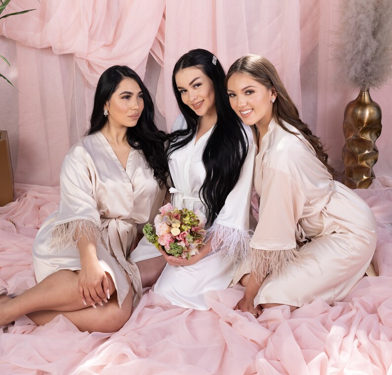 May include: Three women wearing satin robes with feather trim. The robes are in shades of white and blush pink. The women are smiling and looking at the camera. One woman is holding a bouquet of flowers.