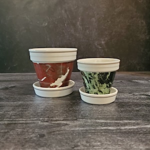 May include: Two ceramic plant pots with white rims and different marbled patterns. The larger pot has a red, white, and black marbled pattern. The smaller pot has a green, black, and white marbled pattern.