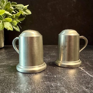 May include: Two vintage pewter salt and pepper shakers. Each shaker is shaped like a small beer stein with a handle and a lid. The shakers have a classic, antique look and are made of a dull, silver-colored metal. The shakers are on a dark surface.