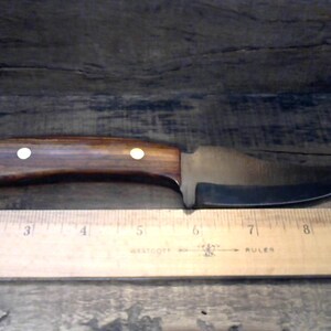 May include: A brown wooden handled knife with a curved blade. The blade is made of metal and has a polished finish. The handle has two white dots on it. The knife is lying on a wooden surface next to a ruler.
