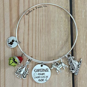 Könnte beinhalten: Silbernes Charm-Armband mit Weihnachts-Themen-Anhängern, darunter ein Grinch, ein Weihnachtsbaum, ein Hund und ein Schlitten. Das Armband hat auch einen Anhänger mit dem Text "Christmas perhaps means a little bit more."