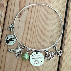Könnte beinhalten: Ein silbernes Armband mit einem grünen Perlenanhänger, einem Frosch-Anhänger, einem Lotusblüten-Anhänger, einem Libellen-Anhänger, einem Schlüssel-Anhänger und einem Anhänger mit dem Text "Mein Traum wäre nicht komplett... ohne dich darin." Das Armband ist mit dem Text "Sparkles and Twine, Co." graviert.
