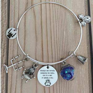 Puede incluir: Pulsera de abalorios plateada con varios colgantes, incluyendo una campana, un tridente, una silla y un globo de cristal lleno de estrellas azules. Un dije redondo presenta el texto "Wizards and witches wherever you dwell, give us a hint, by ringing a bell."