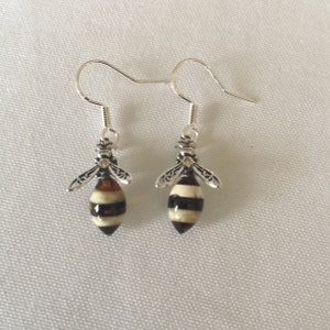 May include: A pair of bee-shaped earrings. Each earring features a silver-colored hook and a bee pendant. The bee pendants have a striped pattern of brown, cream, and dark brown, with silver-colored wings and a head.