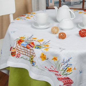 May include: White linen tablecloth featuring embroidered Easter eggs, chicks, and flowers. The tablecloth is laid with a teapot, sugar bowl, and cups.