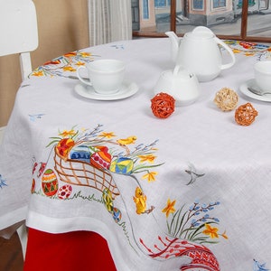 May include: White tablecloth with embroidered Easter eggs, flowers, and birds. A white teapot, sugar bowl, and two cups are set on the table with orange decorative balls.
