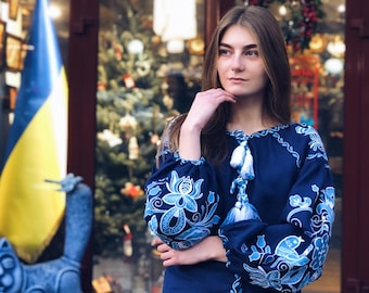 Könnte beinhalten: Eine Frau trägt eine blaue bestickte Bluse mit weißen Blumenmustern und Quasten. Sie steht vor einem Geschäft mit einem Schild, auf dem "Open Ukraine Now" in gelben Buchstaben auf blauem Hintergrund steht.