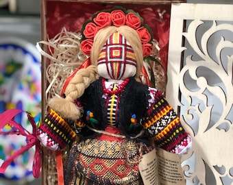 May include: A traditional Ukrainian folk doll, known as a Motanka, dressed in a colorful embroidered outfit with a red flower crown. The doll is handmade and features a unique design with geometric patterns. The doll is displayed in a wooden box with straw.