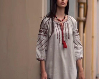 May include: A woman wearing a long, white linen dress with red and black embroidery. She is wearing a red beaded necklace and has long, dark hair. She is holding a black bag.