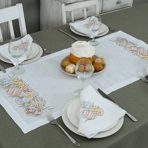 May include: White linen table runner with embroidered Easter eggs in pastel colors. The runner is laid on a green tablecloth and features a centerpiece of a sweet bread and painted eggs.