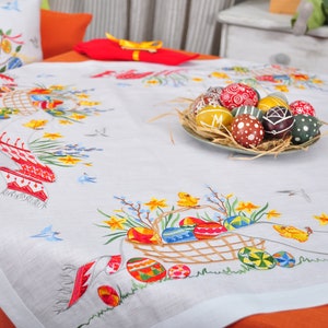 May include: White linen tablecloth with embroidered Easter eggs, flowers, and chicks. A plate of decorated Easter eggs sits in the centre of the table.