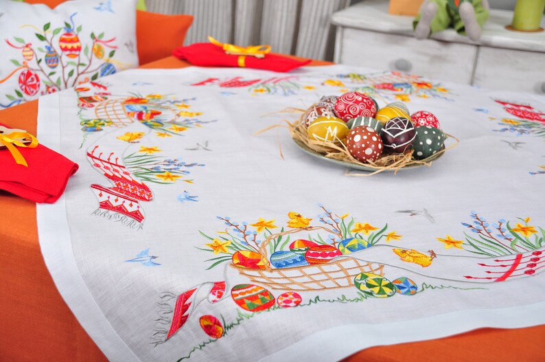 May include: White linen tablecloth with embroidered Easter eggs, flowers, and chicks. A plate of decorated Easter eggs sits in the centre of the table.