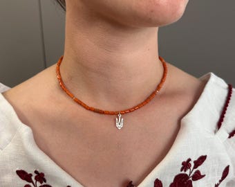 May include: A necklace featuring small, orange beads and a silver pendant in the shape of a trident. The necklace is worn against a white blouse with red floral embroidery. The beads are approximately 0.2 cm in diameter.
