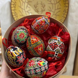Peut inclure: Six œufs de Pâques peints de manière complexe dans une boîte ronde en bois avec du papier de soie rouge. Les œufs sont décorés de motifs géométriques en rouge, noir, vert et blanc.