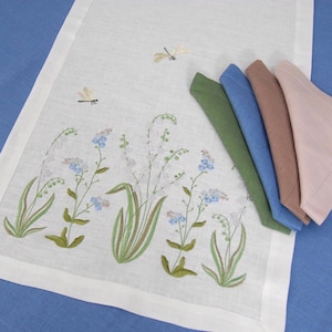 May include: White linen table runner with embroidered floral design featuring white bell flowers, blue forget-me-nots, and green leaves. Two dragonfly designs are also embroidered on the runner. The runner is displayed with four coordinating napkins in green, blue, brown, and pink.