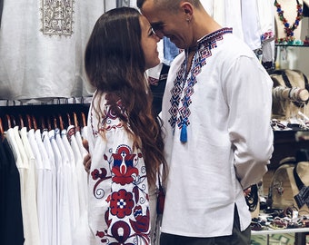 May include: A couple wearing traditional embroidered white shirts with colourful floral patterns. They are standing close together, looking at each other.