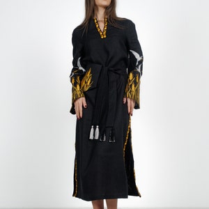 May include: A black linen dress with embroidered yellow wheat designs on the sleeves and a black sash with white tassels. The dress is knee-length and has a V-neckline.