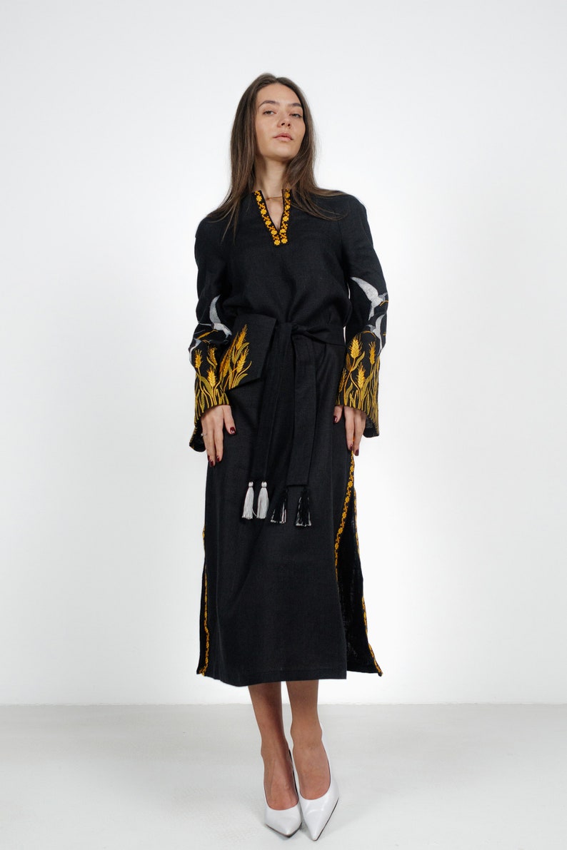 May include: A black linen dress with embroidered yellow wheat designs on the sleeves and a black sash with white tassels. The dress is knee-length and has a V-neckline.