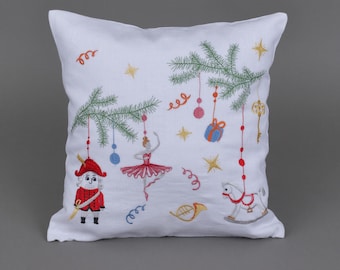 Può includere: Cuscino in lino bianco con decorazioni natalizie ricamate, tra cui uno schiaccianoci, una ballerina, un cavallo a dondolo, un corno musicale e un albero di Natale con ornamenti.