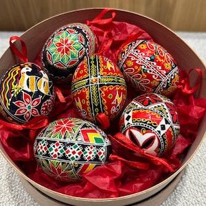Può includere: Sei uova di Pasqua dipinte a mano in una scatola rotonda di legno con carta velina rossa. Le uova sono decorate con intricati motivi geometrici in rosso, nero, verde e giallo.