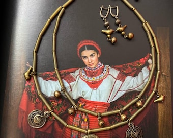 May include: A gold-toned necklace and earrings set. The necklace features a crescent pendant, spiral charms, and bird-shaped accents. The earrings have bird and bell charms. The jewelry is displayed on a dark background with a woman in traditional clothing.