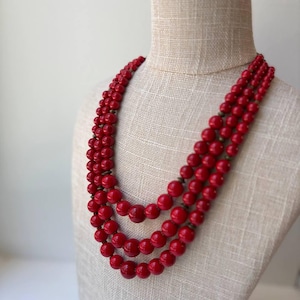 May include: A three-strand red beaded necklace displayed on a mannequin. The beads are round and have a glossy finish. The necklace is a vibrant shade of red and appears to be vintage.