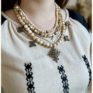 May include: A layered necklace with multiple strands of cream-colored shell beads and a large, ornate, gold-toned cross pendant.