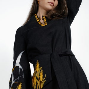 May include: A black linen dress with yellow embroidered wheat stalks and white birds on the sleeves. The dress has a tie belt and a v-neck.