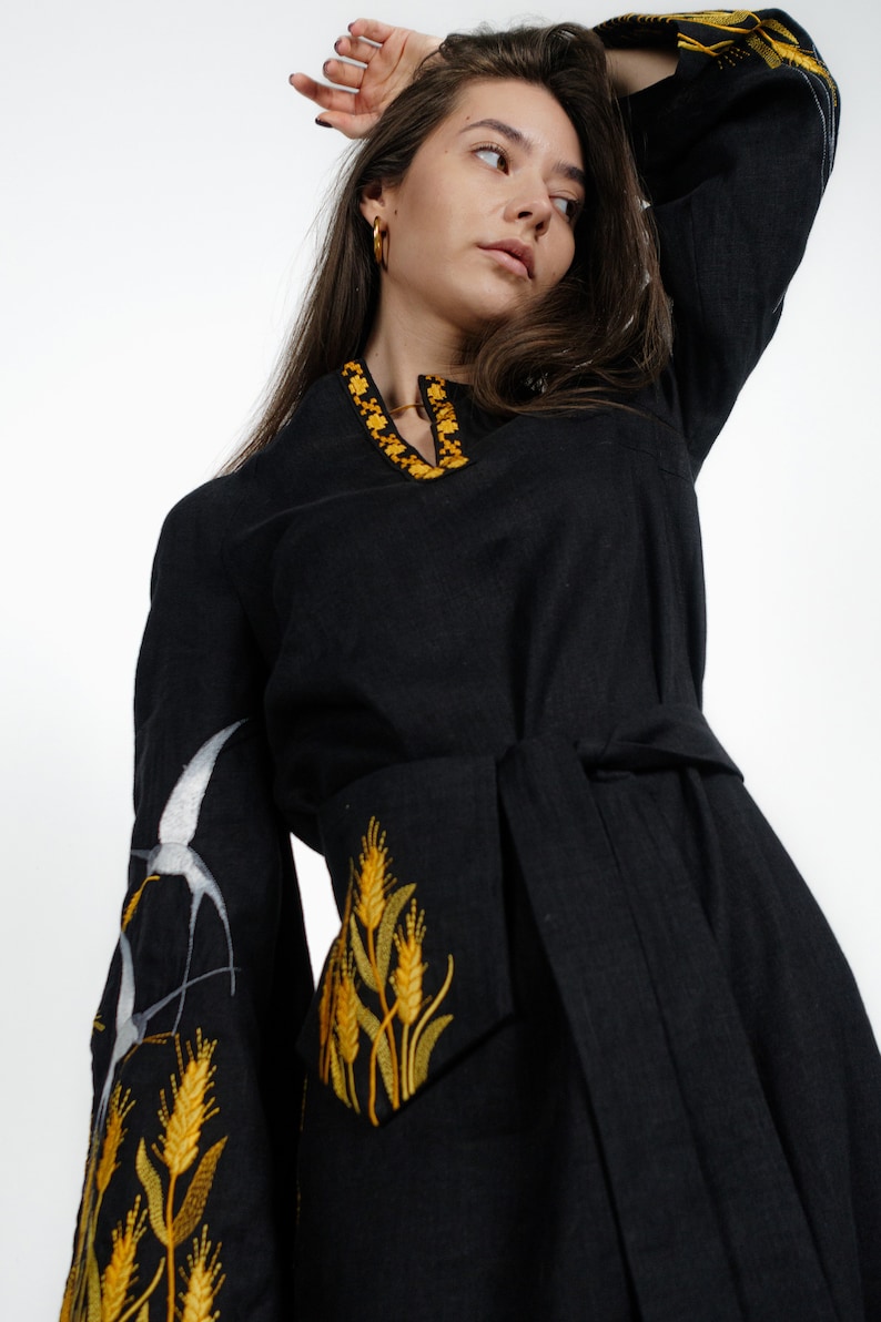 May include: A black linen dress with yellow embroidered wheat stalks and white birds on the sleeves. The dress has a tie belt and a v-neck.
