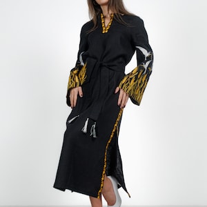 May include: A black linen dress with embroidered white birds and yellow wheat on the sleeves and hem. The dress has a belt and a side slit.