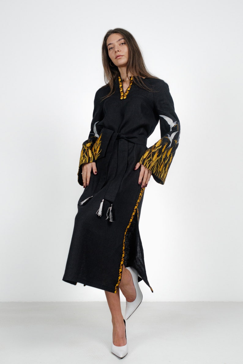 May include: A black linen dress with embroidered white birds and yellow wheat on the sleeves and hem. The dress has a belt and a side slit.