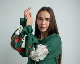 May include: Emerald green tunic with floral embroidery on the sleeves. The sleeves feature red and white floral patterns and white tassels. The tunic has a round neckline and long, gathered sleeves.