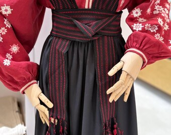 Pode incluir: Uma blusa vermelha de manga comprida com bordados geométricos brancos, combinada com uma saia preta lisa e uma faixa larga tecida em preto com riscas vermelhas finas e longas franjas pendentes em vermelho e preto nas pontas.