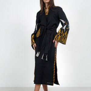 May include: A black linen dress with embroidered yellow wheat and white birds. The dress has a black sash and white tassels. The model is wearing white heels.