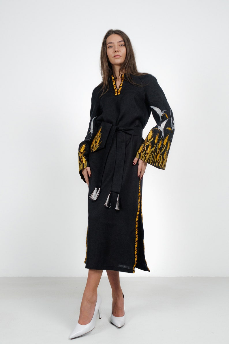 May include: A black linen dress with embroidered yellow wheat and white birds. The dress has a black sash and white tassels. The model is wearing white heels.