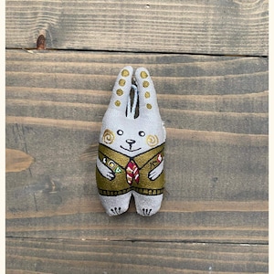 May include: A grey fabric bunny with gold accents and a brown and red tie. The bunny is wearing a green jacket with gold details.