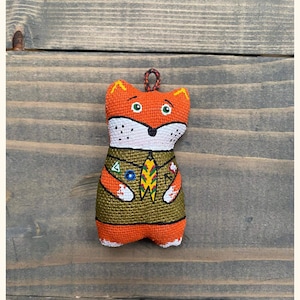 May include: A handmade orange fox plush toy wearing a green and gold uniform with badges. The fox has a white face with black eyes and a black nose.