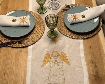 Può includere: Una tavola apparecchiata con un runner in lino con un ricamo a forma di angelo. L'allestimento comprende piatti blu, sottopiatti intrecciati, calici verdi e tovaglioli con anelli a forma di stella. È presente anche un candelabro di vetro con una candela accesa.