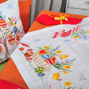 May include: White embroidered tablecloth with an Easter theme. The tablecloth features a basket of colourful eggs, daffodils, and chicks. There is also a pillow with an embroidered Easter tree with colourful eggs.