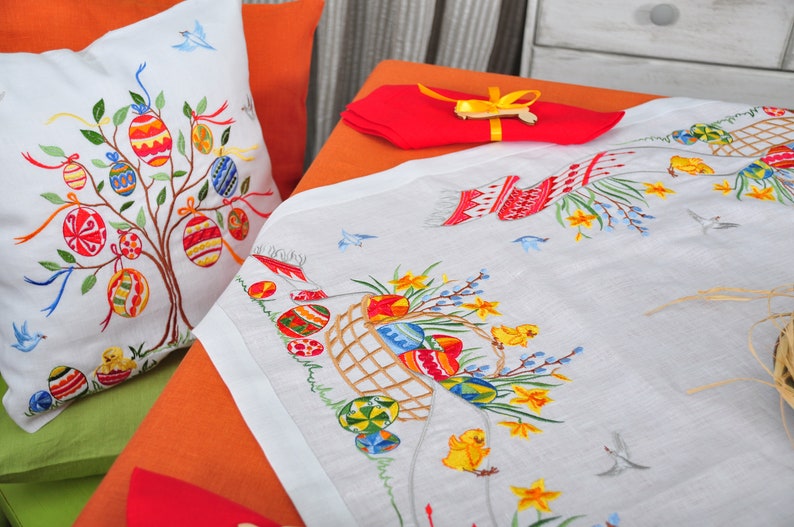May include: White embroidered tablecloth with an Easter theme. The tablecloth features a basket of colourful eggs, daffodils, and chicks. There is also a pillow with an embroidered Easter tree with colourful eggs.