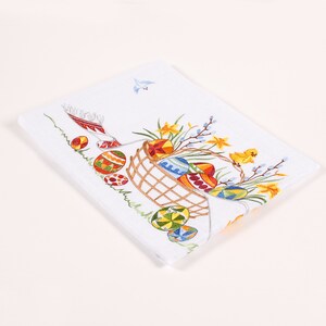 May include: White embroidered kitchen towel featuring a basket overflowing with colourful Easter eggs, yellow daffodils, and a small yellow chick. The basket is surrounded by green grass and a few more eggs.