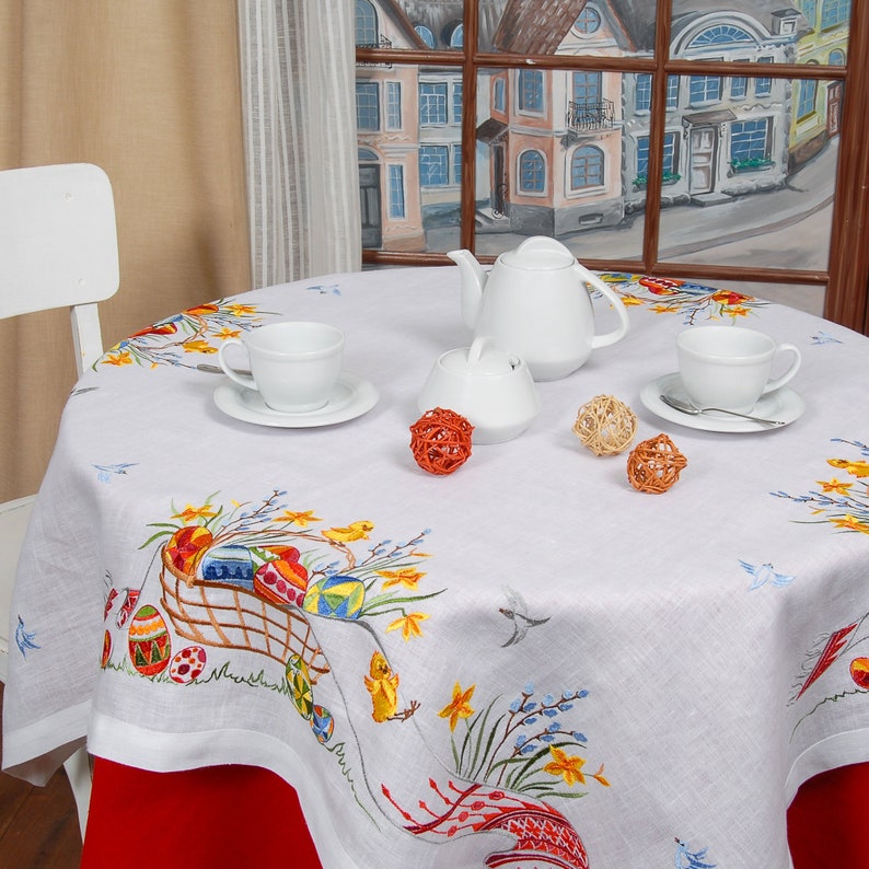 May include: White tablecloth with embroidered Easter eggs, flowers, and birds. A white teapot, sugar bowl, and two cups are set on the table with orange decorative balls.