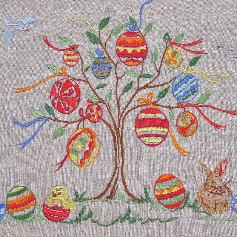 K&ouml;nnte beinhalten: Ein gestickter Osterbaum mit bunten Eiern und einem Hasen. Der Baum hat braune &Auml;ste und gr&uuml;ne Bl&auml;tter. Die Eier sind mit Streifen, Punkten und anderen Mustern verziert. Der Hase ist braun und wei&szlig; und sitzt auf dem Boden.