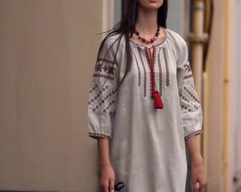 May include: A woman wearing a long, white linen dress with red embroidery and a red beaded necklace. She is walking down a street and carrying a black bag.